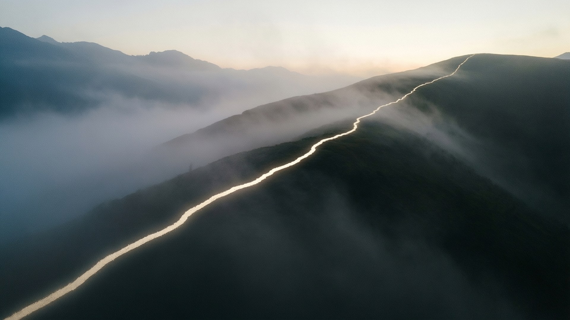 Deep Meaning Lines 桌布 壁紙 — 一條蜿蜒的道路穿過霧氣瀰漫的山脈，在黎明時分，柔和的金色光芒照亮了雲海之上的路徑。