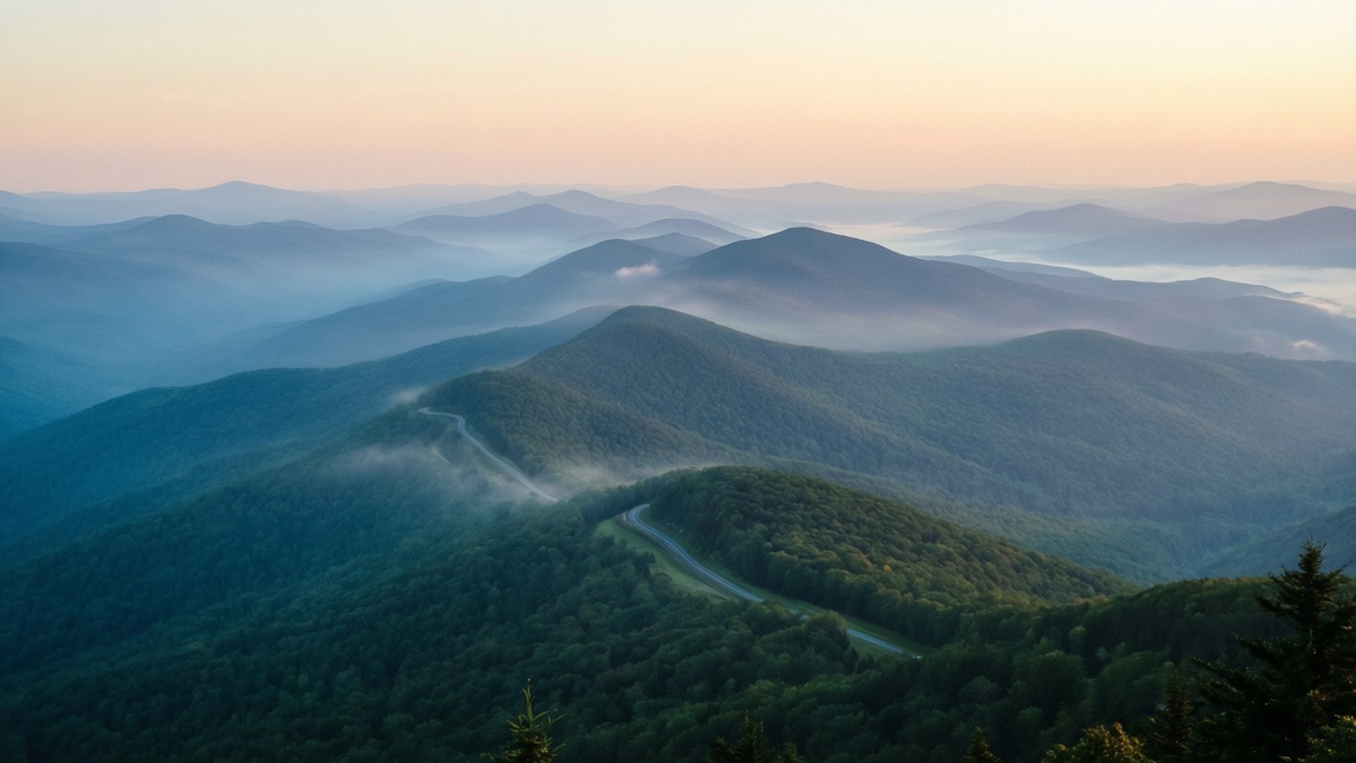 Misty Blue Ridge Morning 壁紙 — 一條蜿蜒的道路在黎明時分蜿蜒穿過霧氣瀰漫的森林山脈，天空呈現柔和的粉紅色和藍色色調。