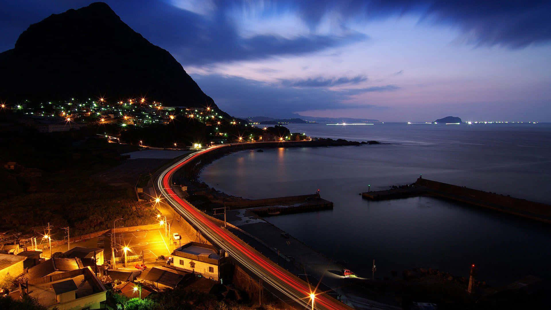 壁紙 — 一條蜿蜒的海岸公路，在暮色中帶有汽車的光跡、發光的村莊燈光，以及紫色天空下的黑暗山脈輪廓。