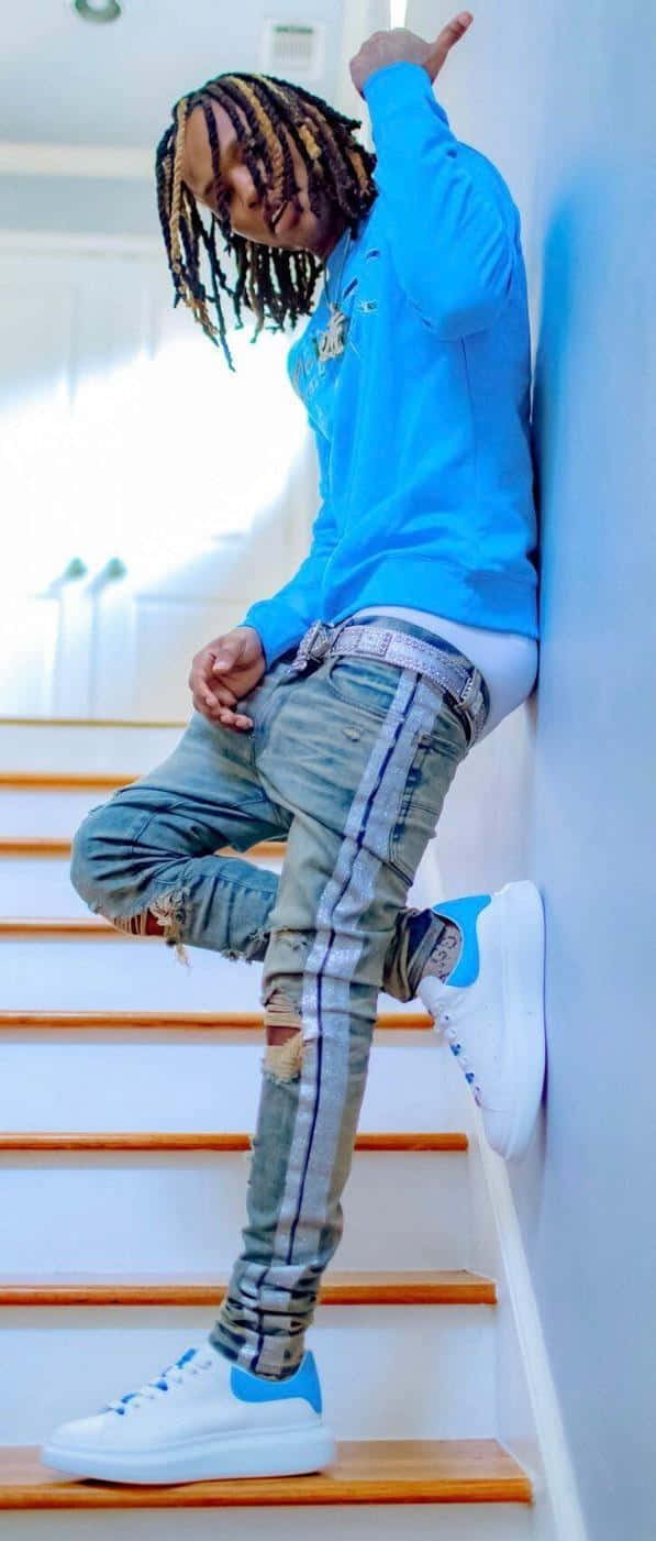  壁紙 — 一位辮子髮型的年輕男子隨意地靠在木樓梯旁的牆上，穿著亮藍色運動衫和破洞牛仔褲。