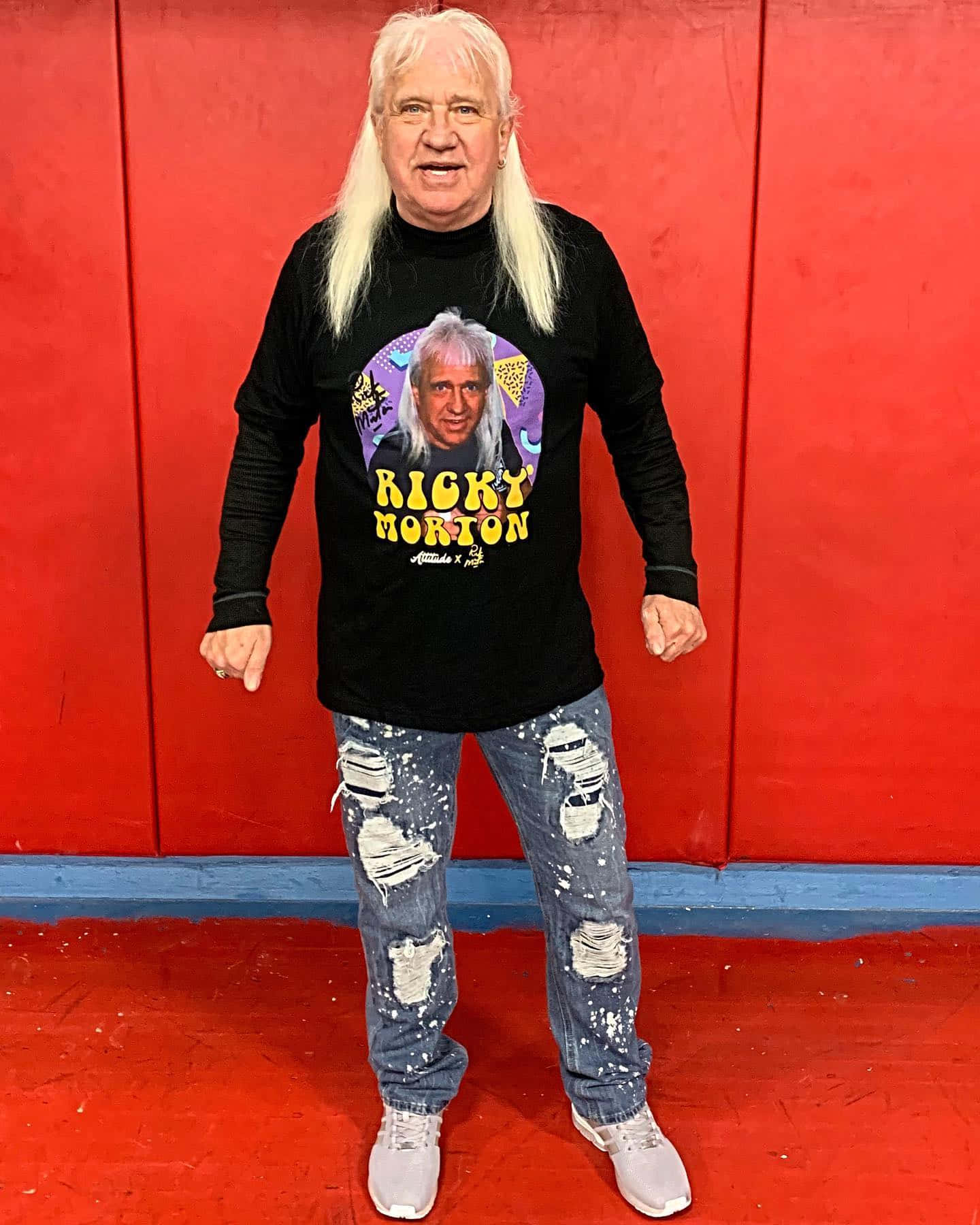  壁紙 — 一位長白髮的男子穿著黑色Ricky Morton T恤和破洞牛仔褲，站在大膽的紅牆前。
