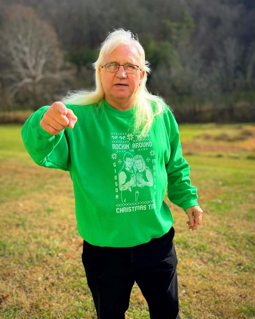  壁紙 — 一位長金髮戴眼鏡的男子，穿著亮綠色聖誕毛衣，在草地上向前指。