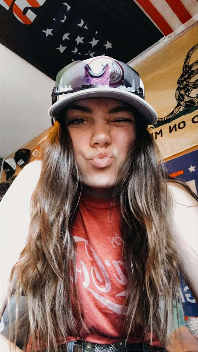  壁紙 — 一位長棕色頭髮的年輕女性在室內戴著帽子、穿著紅色T恤，做出俏皮的嘟嘴表情。