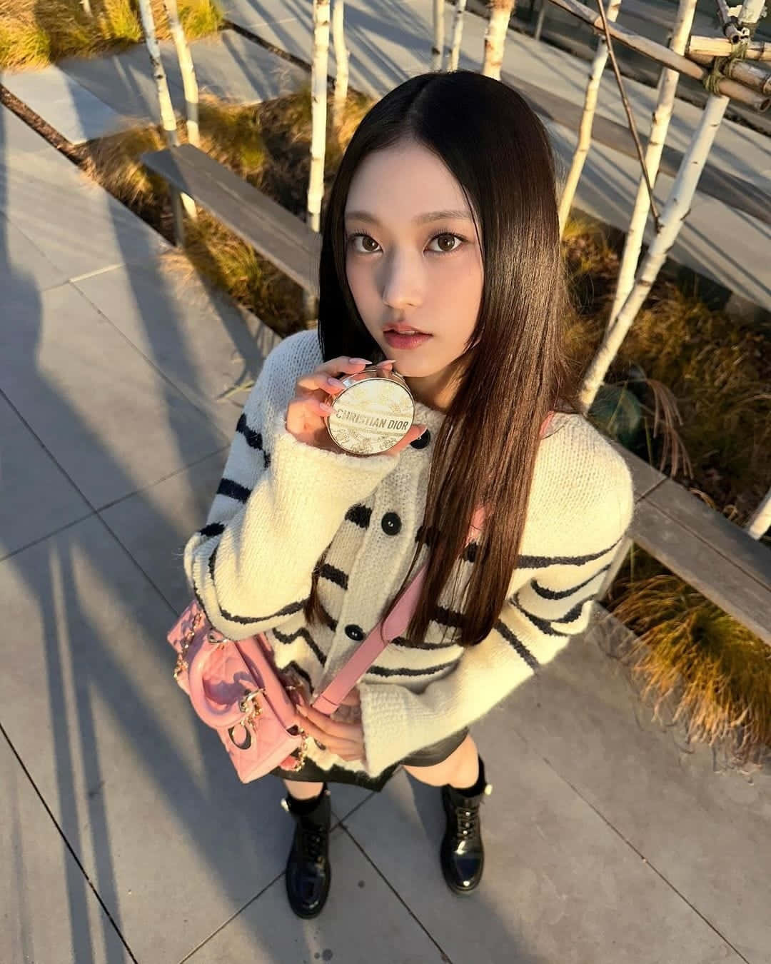  壁紙 — 一位長黑髮的年輕女性站在陽光普照的木甲板上，手持Christian Dior粉餅，身穿米色和黑色條紋毛衣及粉紅色手提包。
