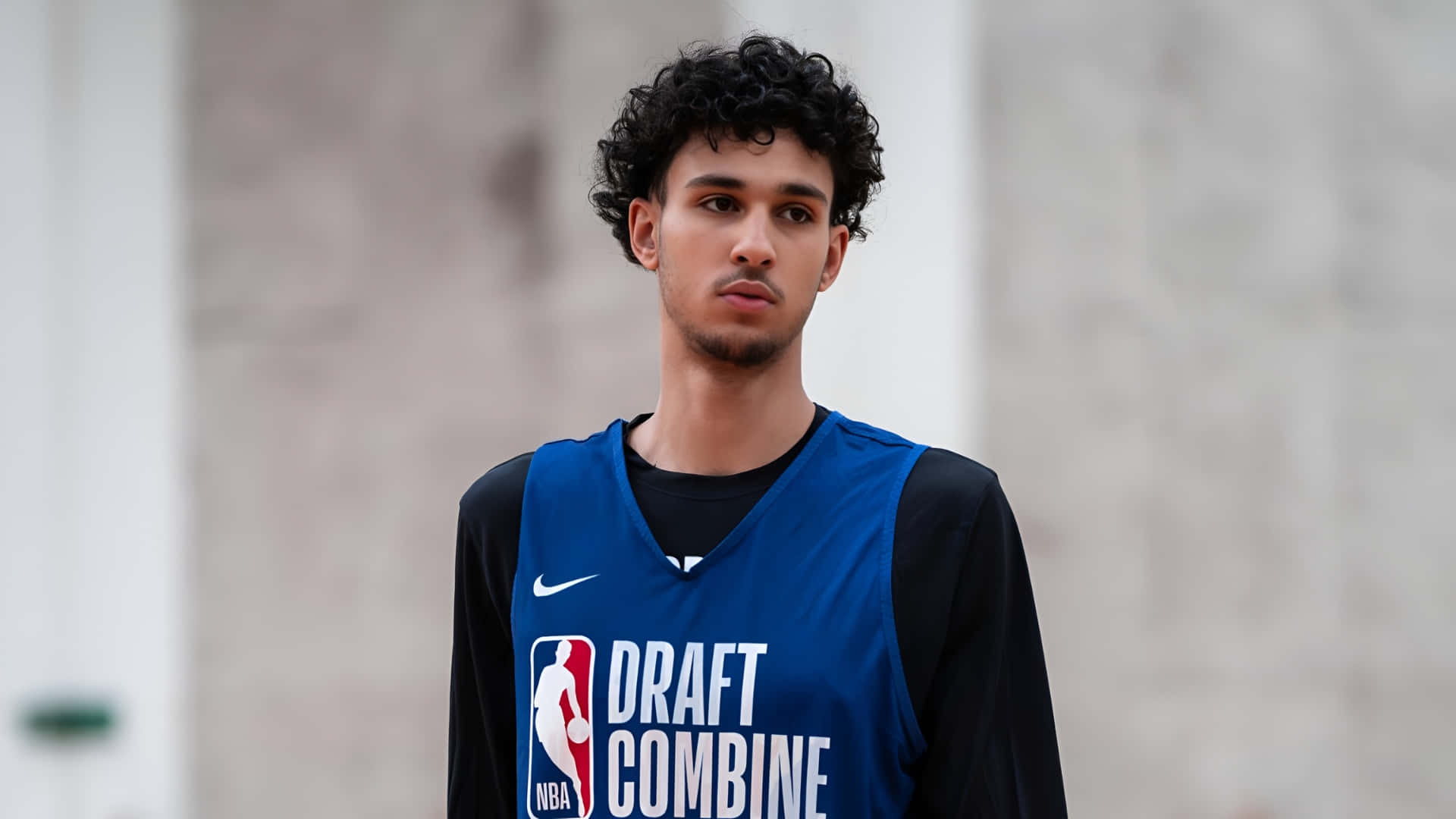  壁紙 — 一位身穿藍色NBA選秀聯合試訓球衣的年輕籃球運動員，在模糊的混凝土牆前帶著專注的表情站立。
