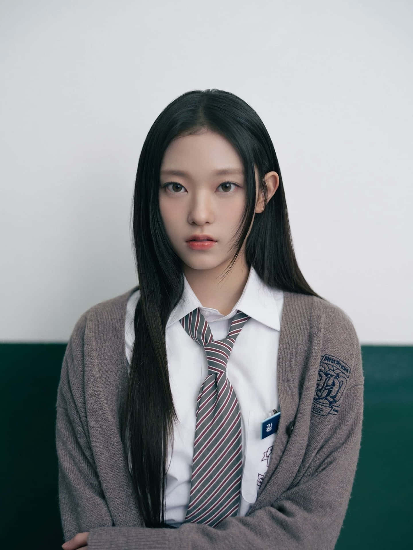  壁紙 — 一位身穿校服、打著條紋領帶和灰色開襟毛衣的年輕女性，靠著雙色牆壁，表情嚴肅地直視鏡頭。