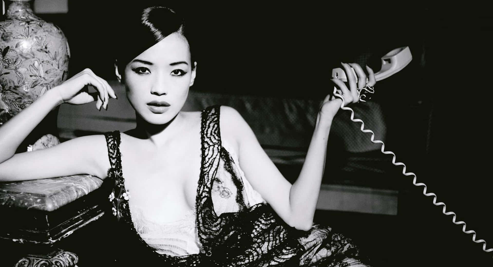 Shu Qi 桌布 壁紙 — 一位穿著蕾絲內衣的優雅女性，在昏暗的房間裡坐在華麗的花瓶旁，手持一部復古電話。