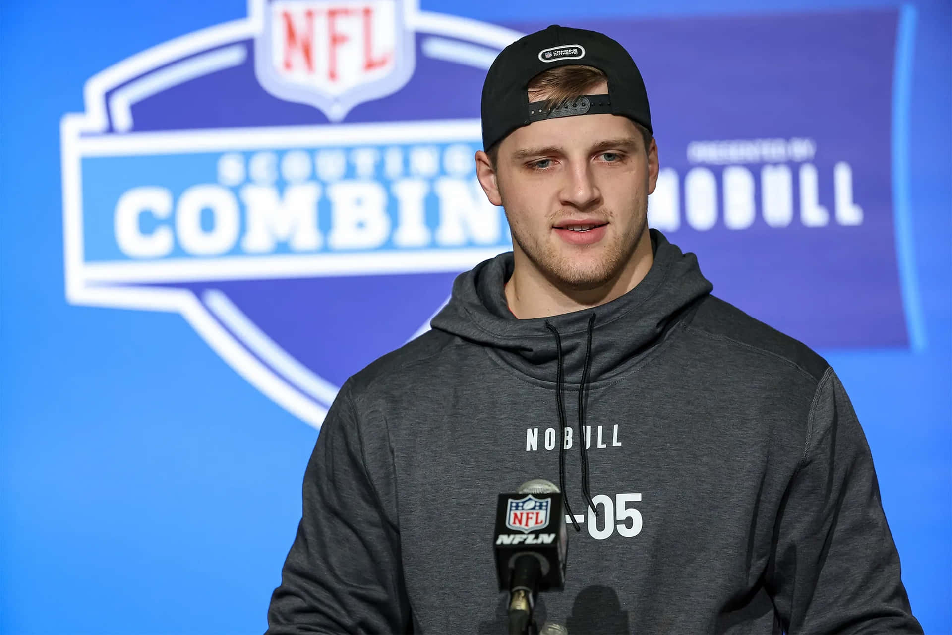  壁紙 — 一位戴黑色帽子和灰色連帽衫的年輕NFL球員在NFL選秀聯合試訓記者會上發言。