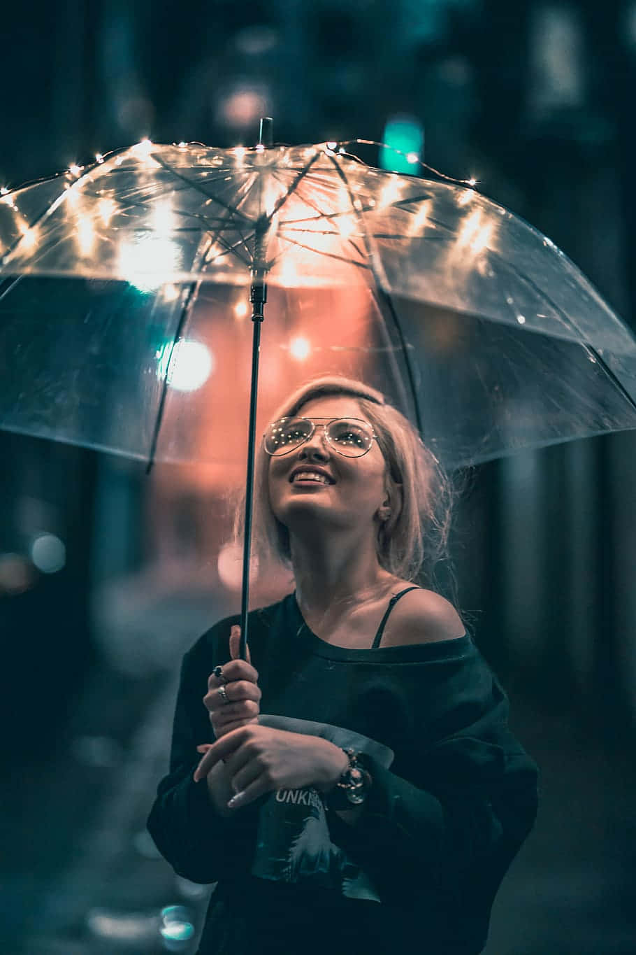  壁紙 — 一位戴眼鏡的微笑女士在模糊的雨夜城市背景下，舉著一把被串燈照亮的透明雨傘。