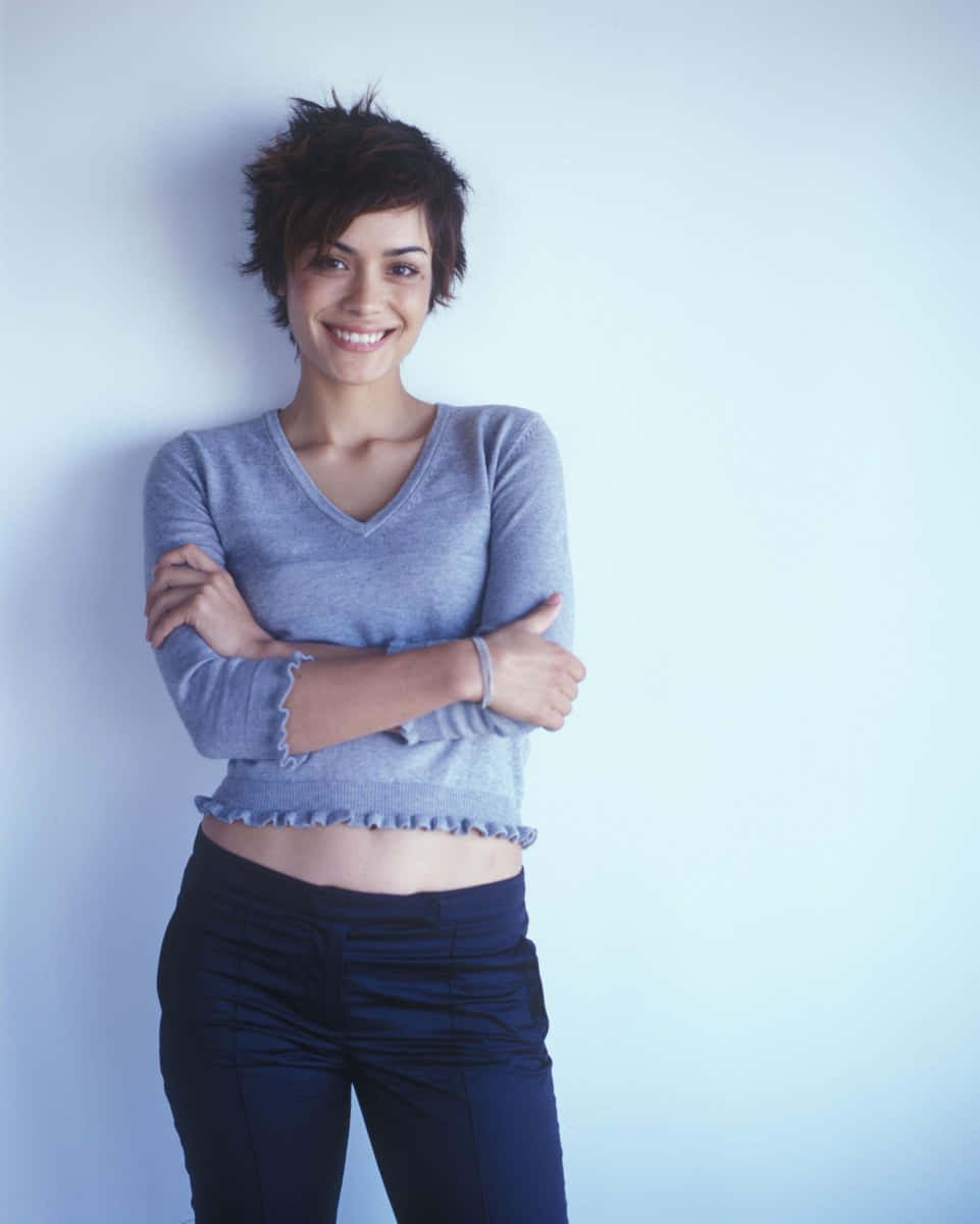 Shannyn Sossamon 桌布 壁紙 — 一位短黑髮微笑的女子靠著淺藍色牆壁，身穿灰色短版毛衣和深色褲子。