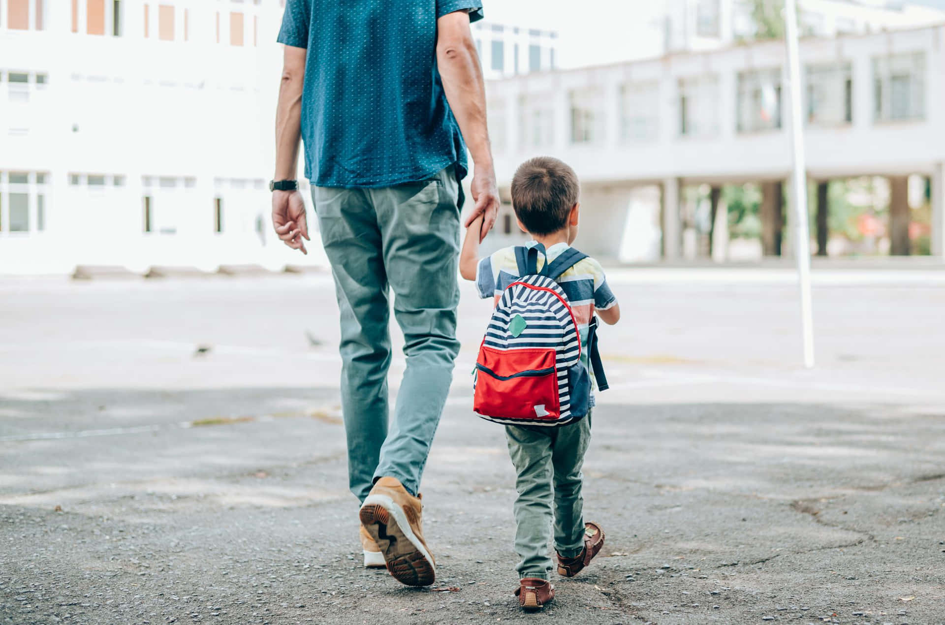  壁紙 — 一位父親和年幼的兒子手牽手走過鋪砌的校園，背著一個色彩繽紛的背包。