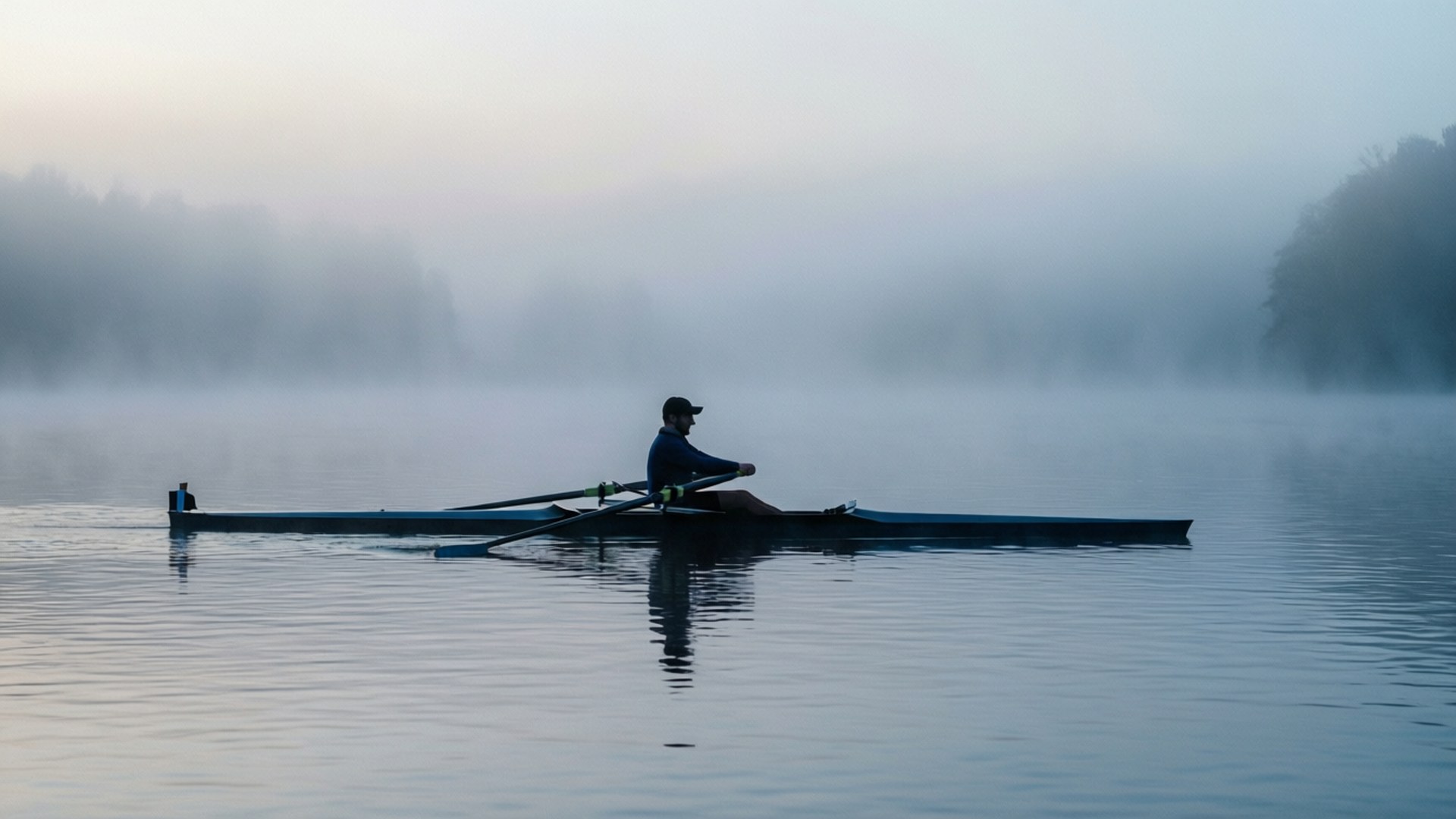 Misty Morning Scull 壁紙 — 一位孤獨的划船者在霧氣瀰漫的黎明水面上靜靜滑行，樹木繁茂的河岸逐漸消失在柔和的灰色薄霧中。