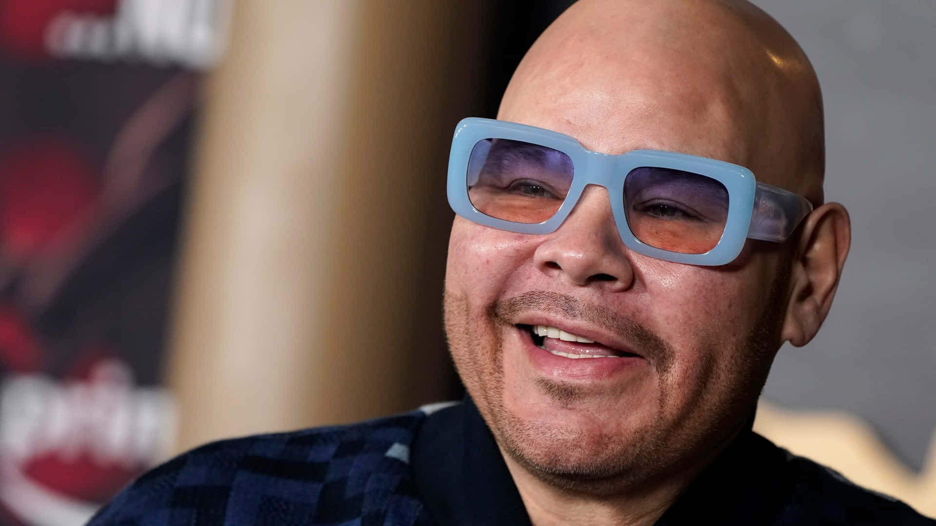  壁紙 — 一位光頭男士帶著溫暖的笑容，在室內柔和燈光下戴著超大淺藍色太陽眼鏡。