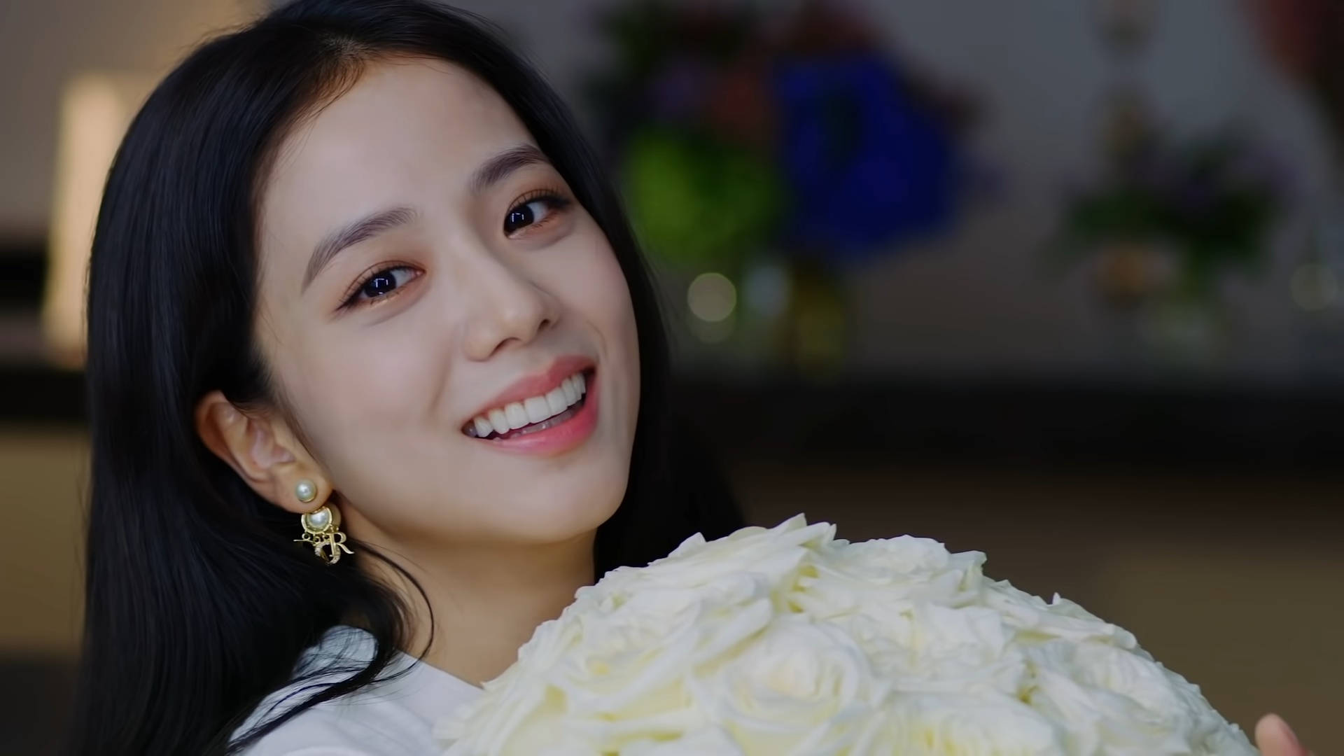 一位微笑的女性在室內手持一束白玫瑰。 — Blackpink 壁紙
