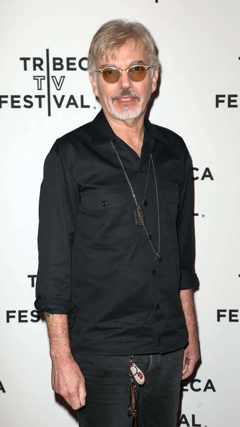  壁紙 — 一位灰髮戴太陽眼鏡的男子，穿著黑色襯衫和牛仔褲，在Tribeca TV Festival活動中對著鏡頭微笑。