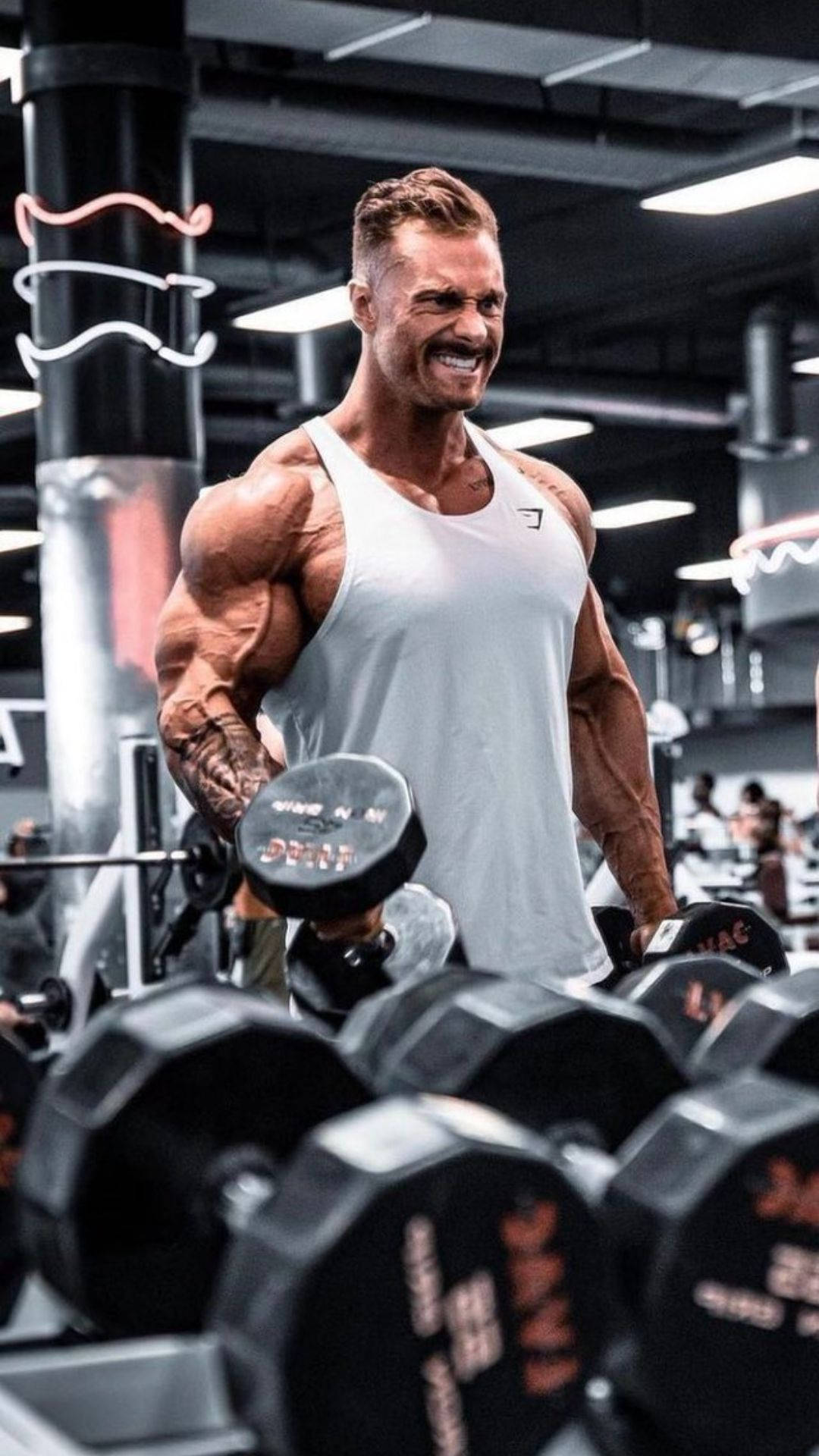 一位微笑的肌肉發達男子在霓虹燈健身房中舉啞鈴。 — Chris Bumstead 壁紙