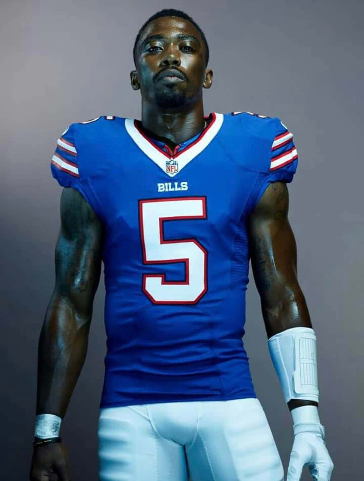  壁紙 — 一位肌肉發達的NFL美式足球員，身穿藍色比爾隊5號球衣，自信地站在純灰色背景前。