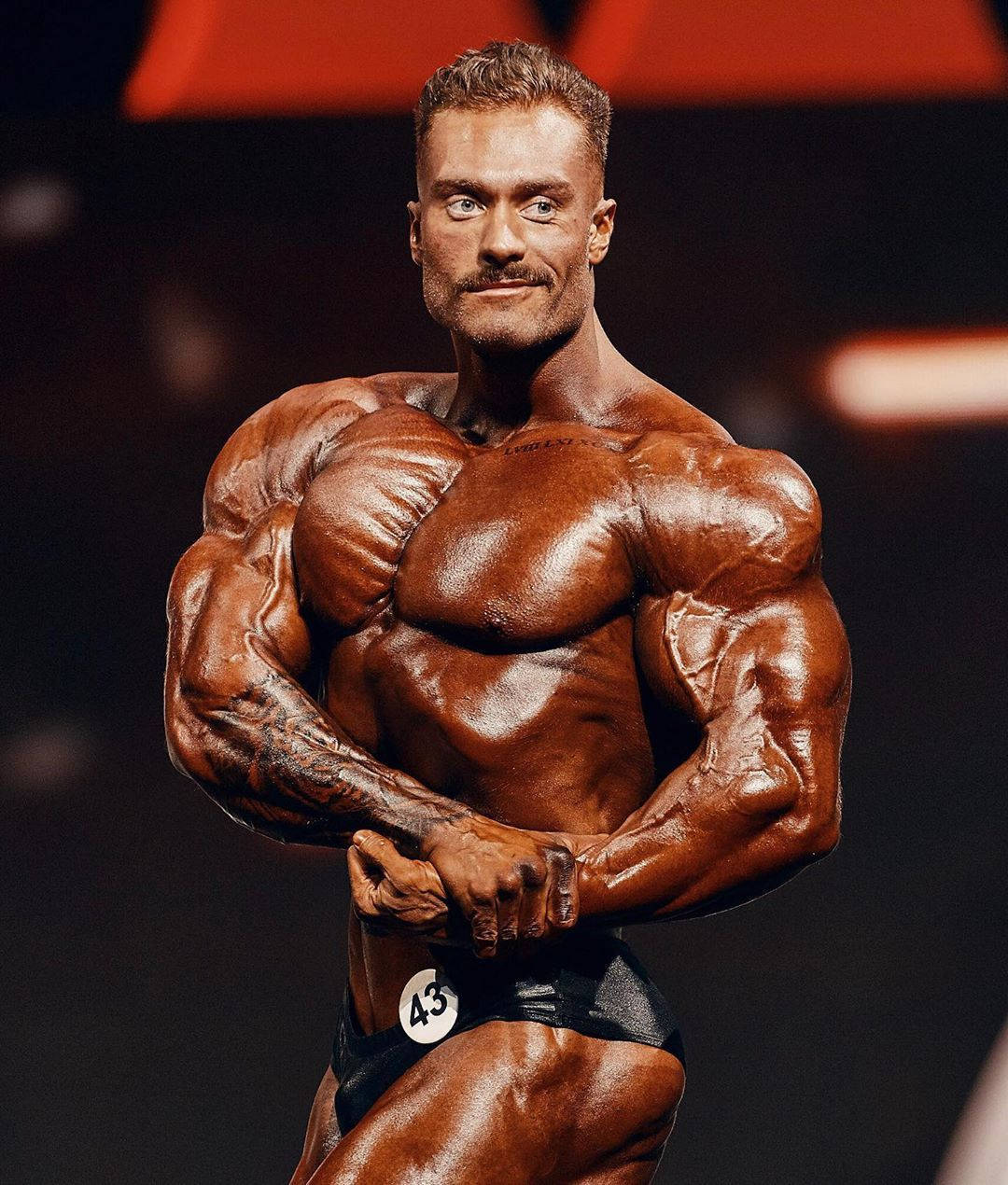 一位男性健美運動員在舞台上擺姿勢，皮膚塗油，比賽號碼為43。 — Chris Bumstead 壁紙