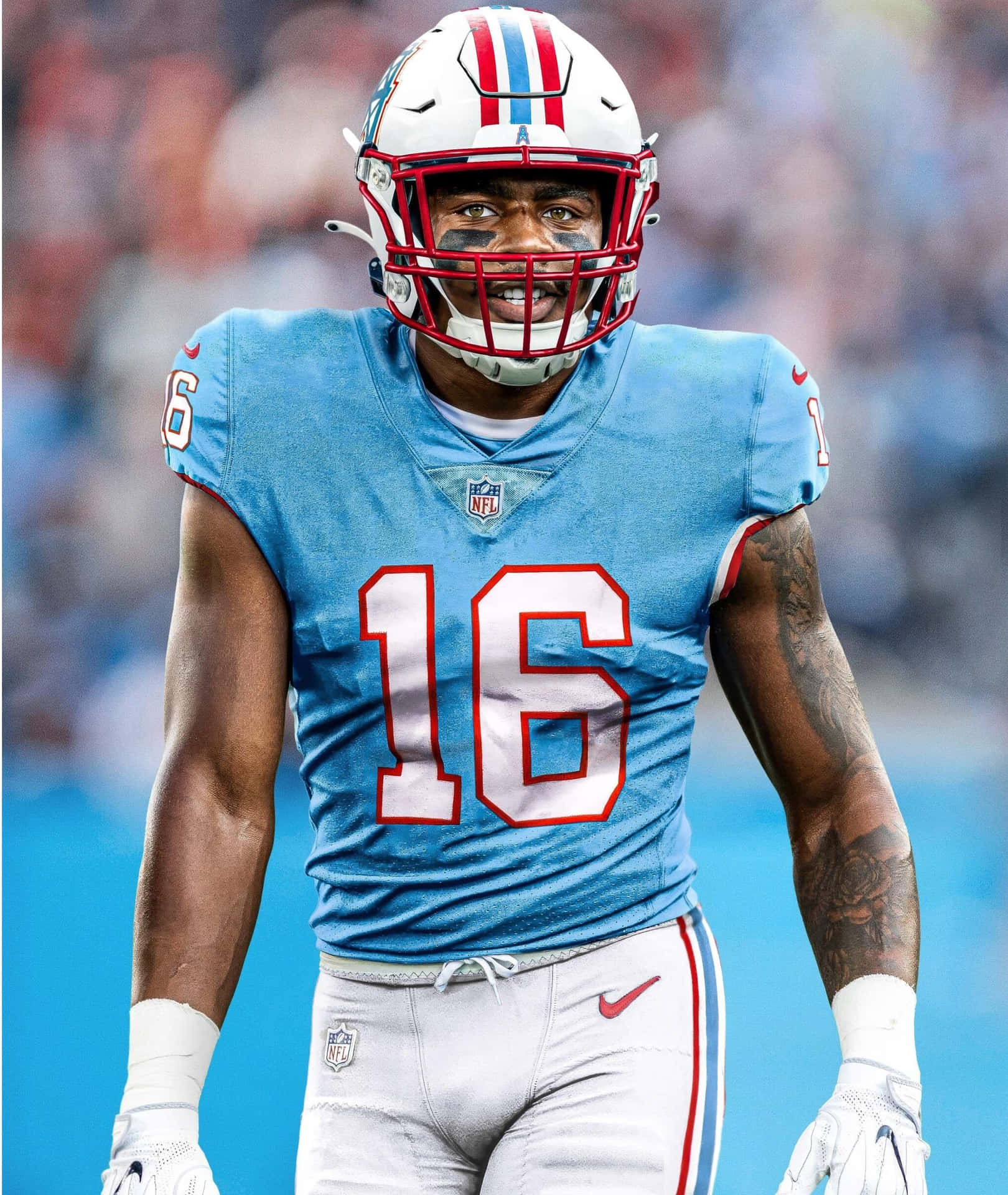  壁紙 — 一位堅定的NFL美式足球員，身穿淺藍色16號球衣，戴著白色頭盔和紅色面罩，手臂上有明顯的刺青，站在球場上。