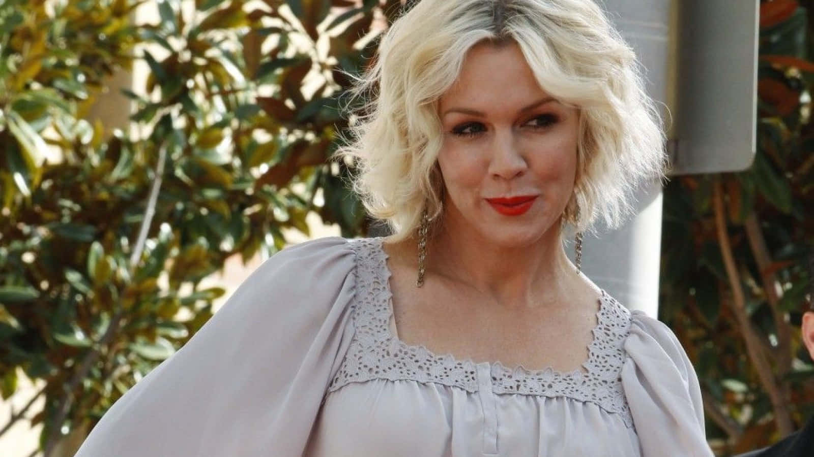 Jennie Garth 桌布 壁紙 — 一位金髮紅唇的女性在戶外穿著一件帶有蕾絲細節的淺灰色襯衫。