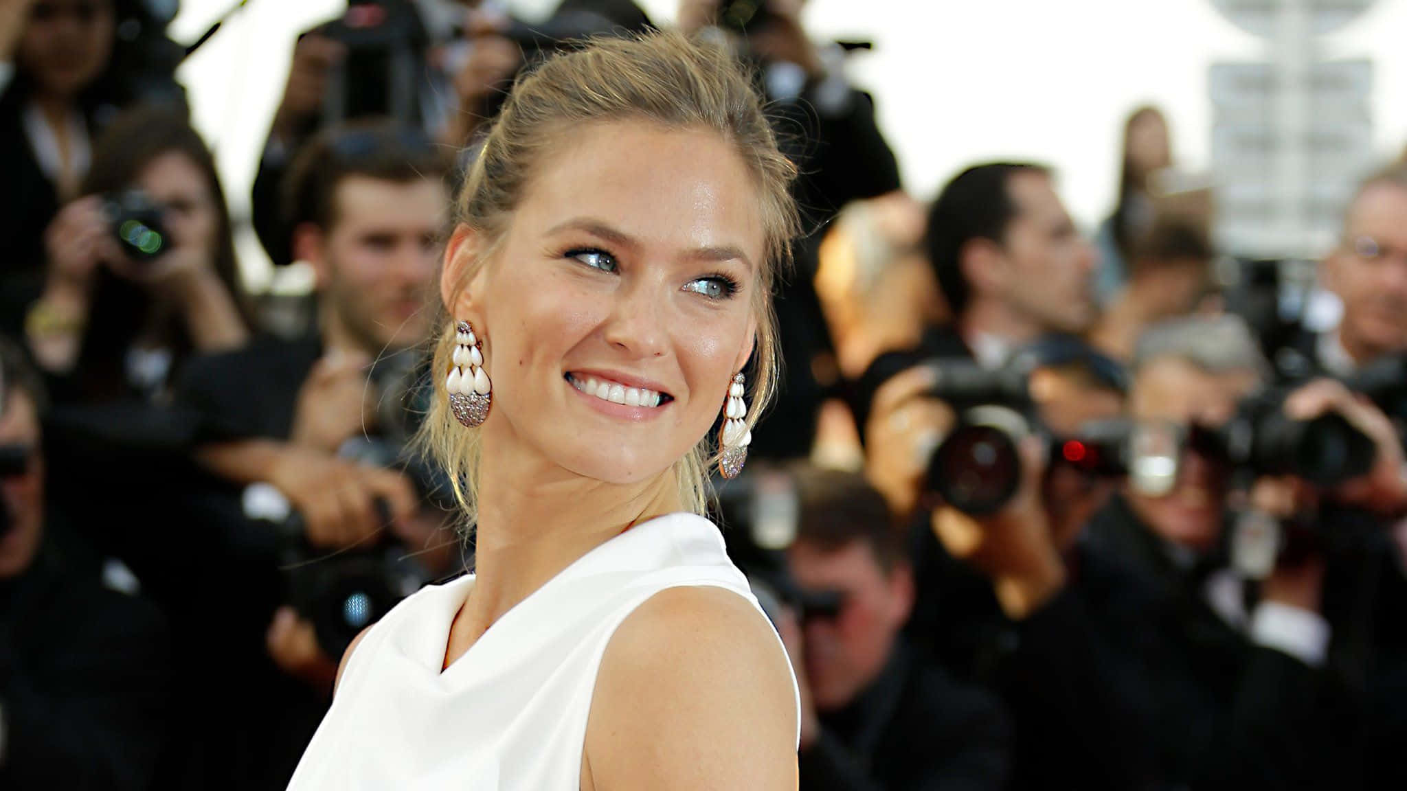 Bar Refaeli 桌布 壁紙 — 一位金髮美女身穿白色連衣裙，在華麗活動中被攝影師包圍，燦爛微笑。
