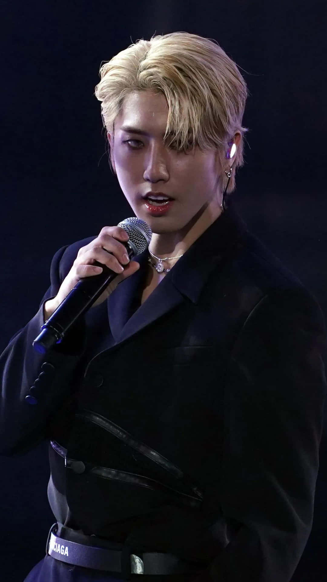  壁紙 — 一位金髮男表演者在舞台燈光下穿著黑色服裝對著麥克風唱歌。