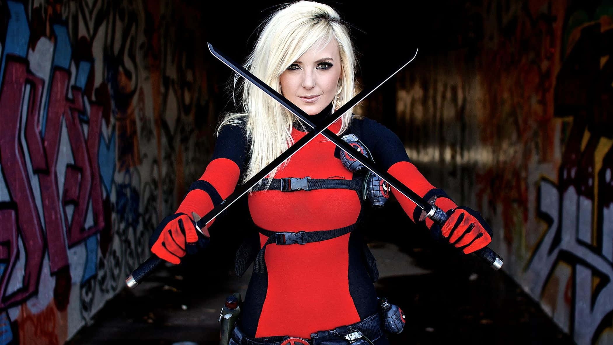 Fantasy Cosplay 桌布 壁紙 — 一位金髮女子身穿紅黑服裝，在佈滿塗鴉的小巷中手持兩把交叉的劍。