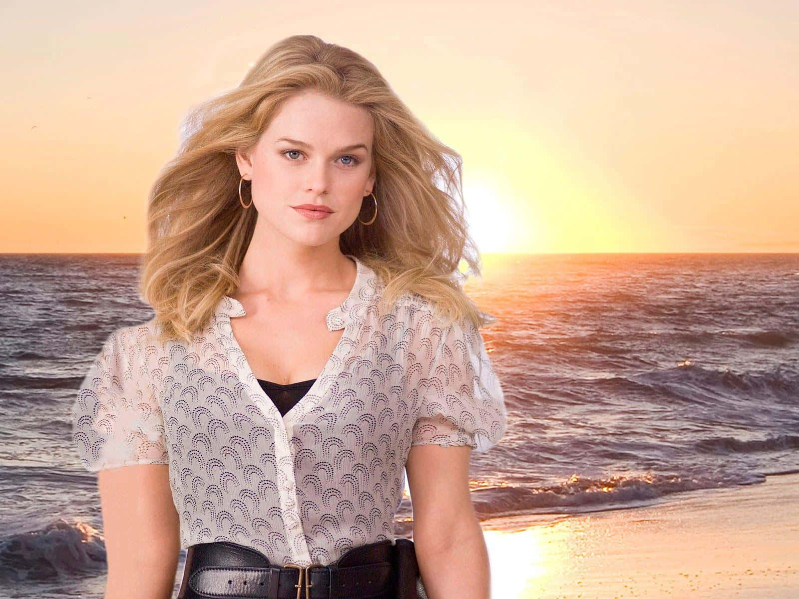 alice eve 桌布 壁紙 — 一位金髮女子在日落時分站在沙灘上，身後有海浪，頭髮上有金色的光。