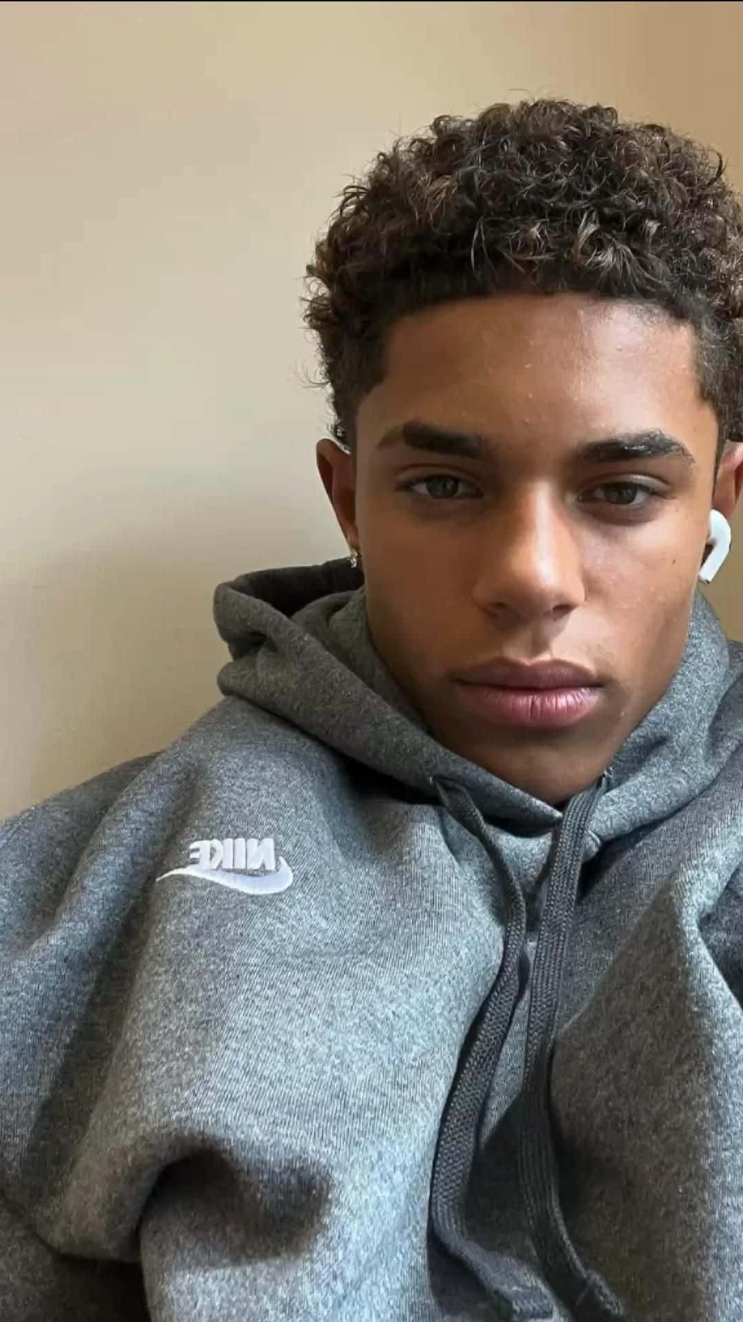  壁紙 — 一位捲髮年輕男子穿著灰色Nike連帽衫和白色耳機，直視鏡頭。