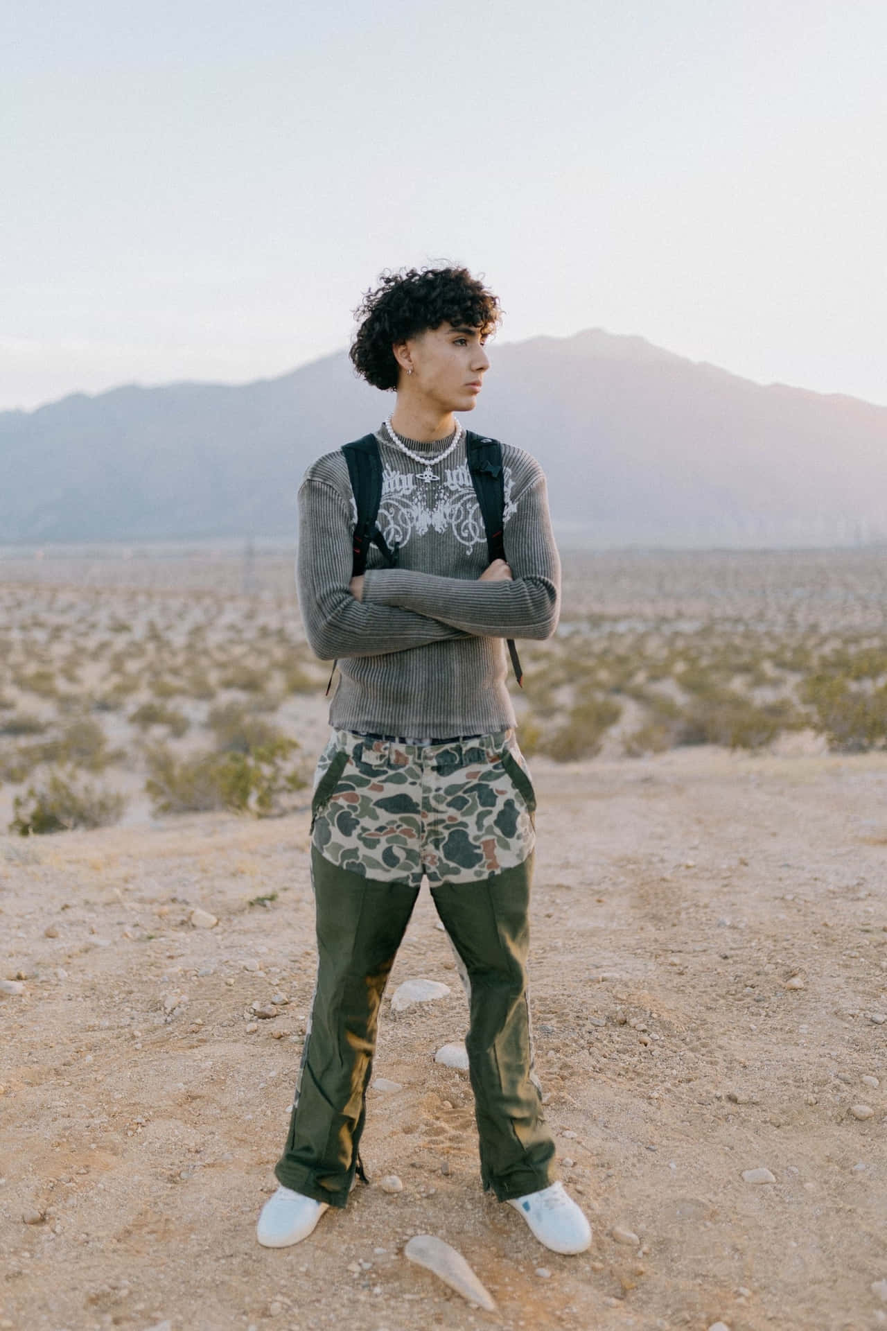  壁紙 — 一位捲髮年輕男子自信地站在沙漠景觀中，雙臂交叉，身穿灰色毛衣和迷彩短褲。