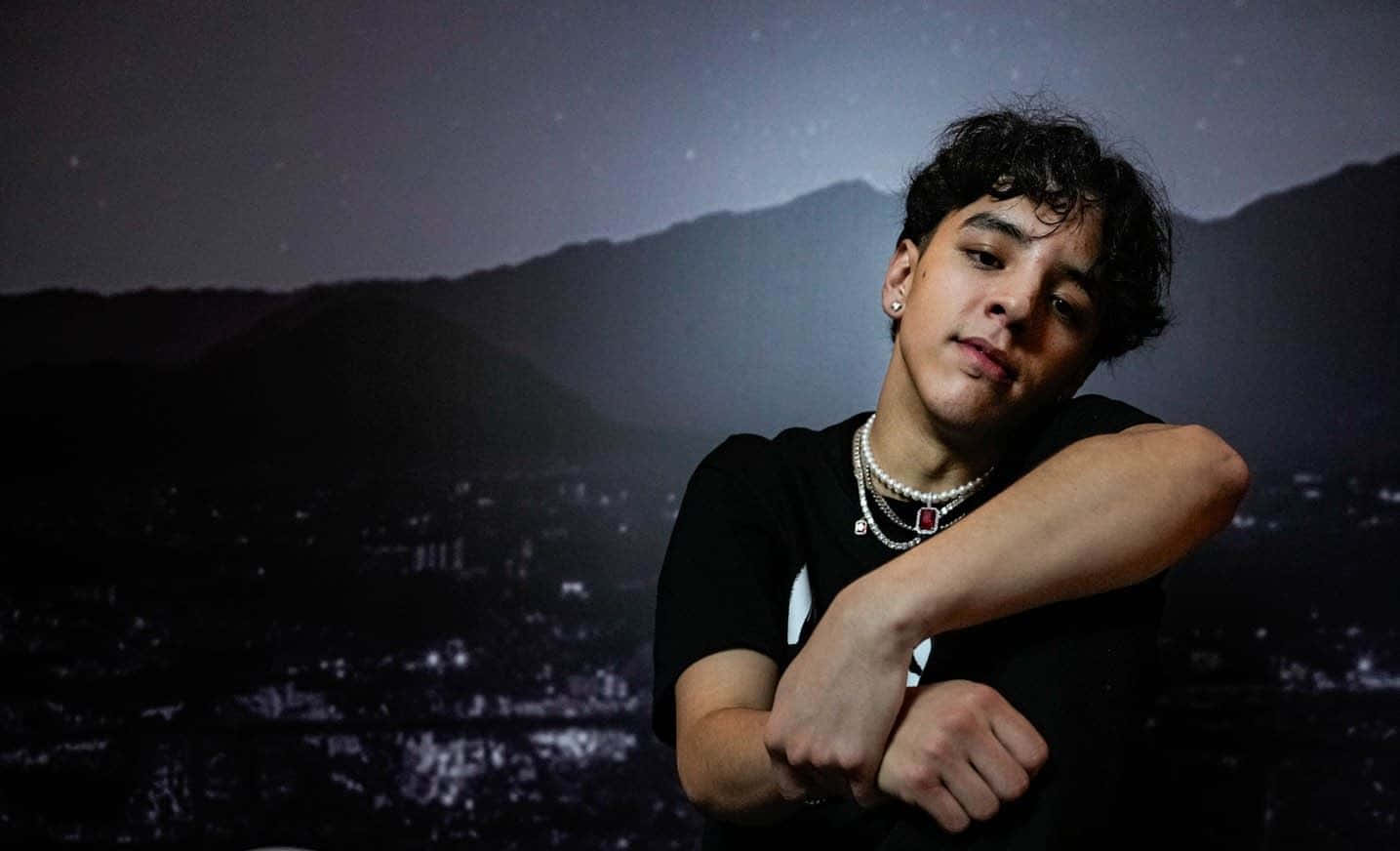  壁紙 — 一位捲髮、佩戴多層項鍊的年輕男子，在星空背景下自信地擺姿勢，下方是剪影般的山脈和城市燈光。