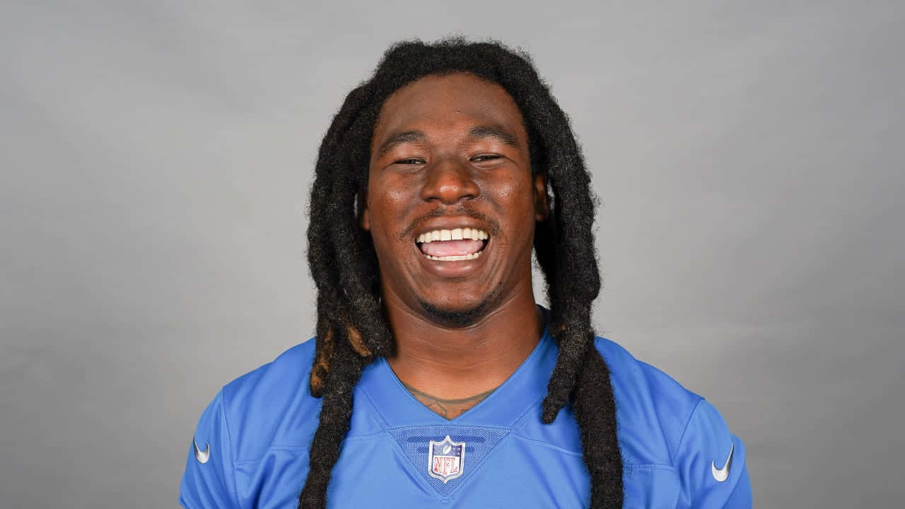  壁紙 — 一位快樂的NFL球員留著長辮子，穿著藍色獅子隊球衣，在純灰色工作室背景前開懷大笑。