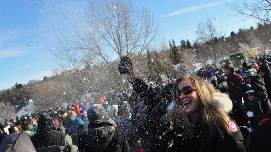  壁紙 — 一位快樂的女人在晴朗藍天下，於擁擠的冬季節慶中笑著將雪拋向空中。