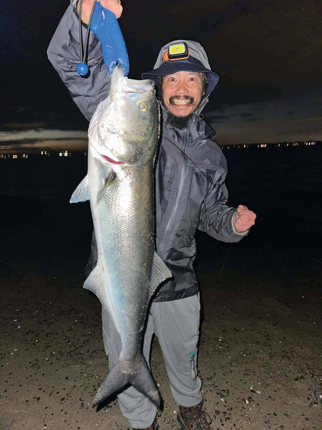  壁紙 — 一位留鬍鬚的漁夫在夜間燈光照耀的海岸線上，自豪地捧著一條巨大的銀色魚，笑容滿面。