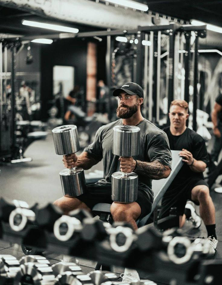 男士在健身房舉啞鈴，另一男士觀看 — Chris Bumstead 壁紙