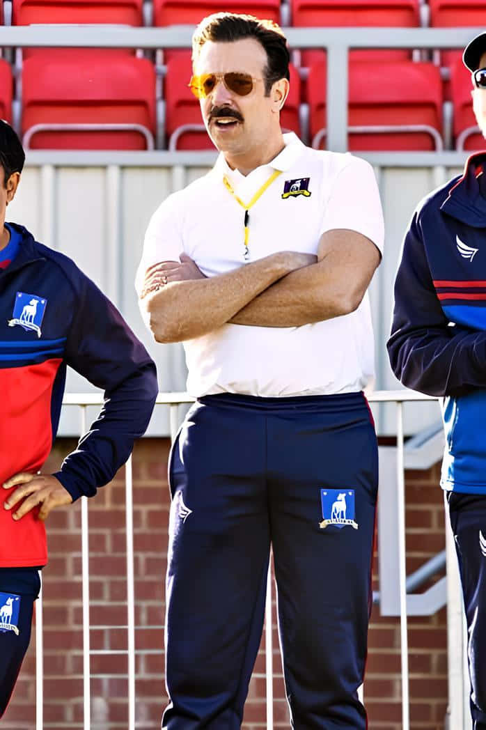  壁紙 — 一位留鬍子、戴太陽眼鏡的男子，身穿白色Polo衫和海軍藍長褲，在體育館內交叉雙臂站立。