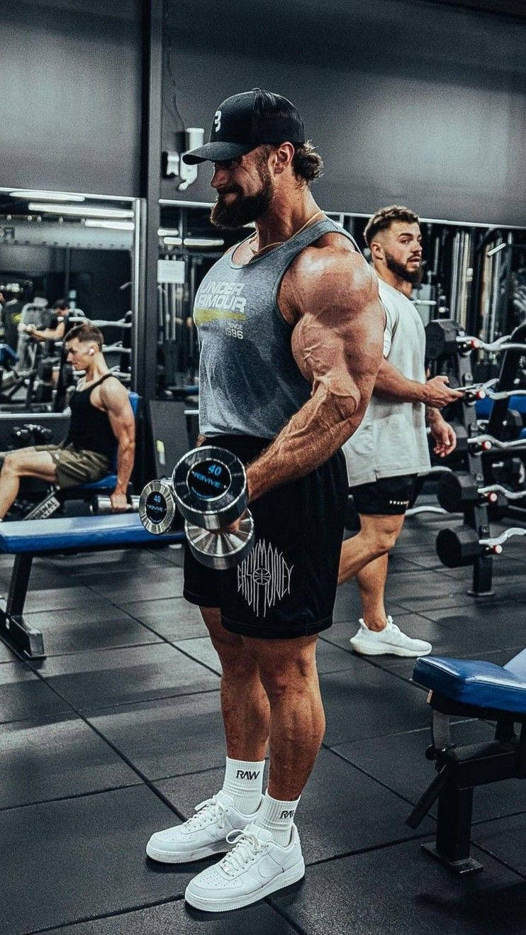 一位肌肉男在健身房舉啞鈴，其他人正在訓練。 — Chris Bumstead 壁紙