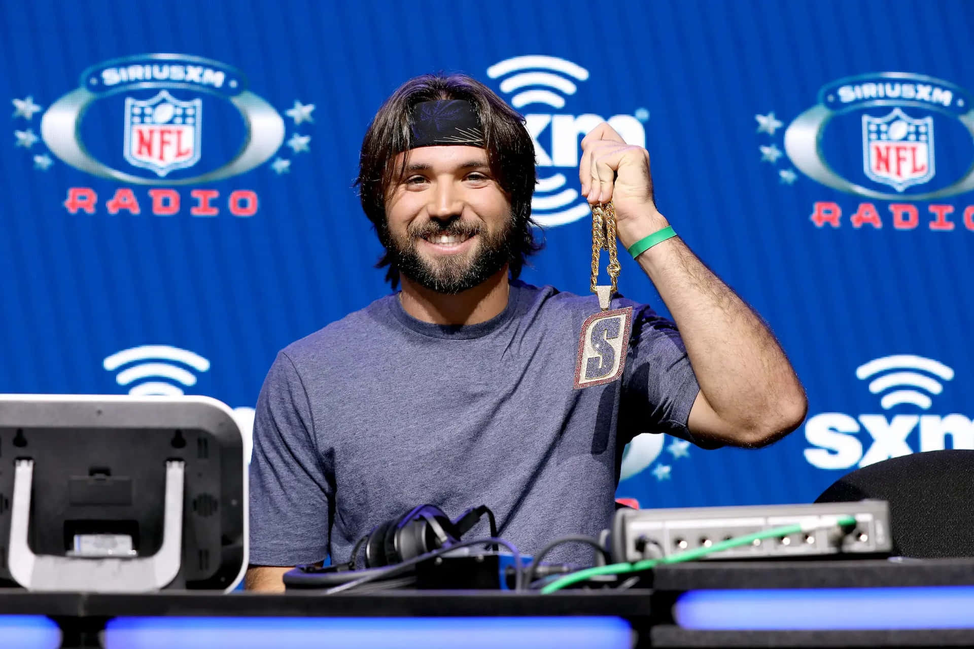  壁紙 — 一位留鬍鬚的微笑男士在藍色 SiriusXM NFL Radio 背景前舉起一個金色 NFL 收音機鑰匙圈。