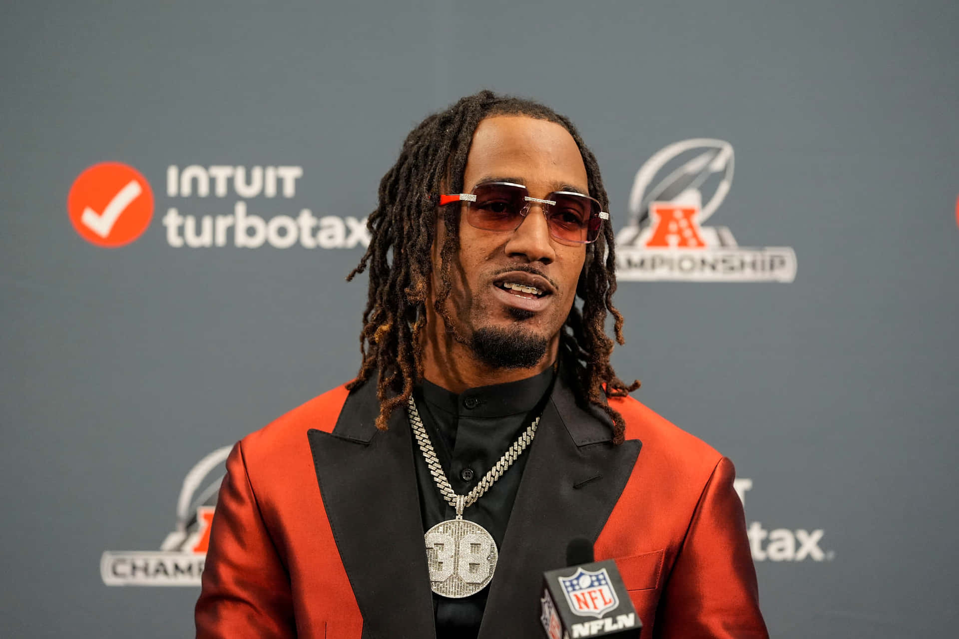  壁紙 — 一位留著辮子頭、戴紅色太陽眼鏡的足球員在記者會上對著NFL麥克風說話。