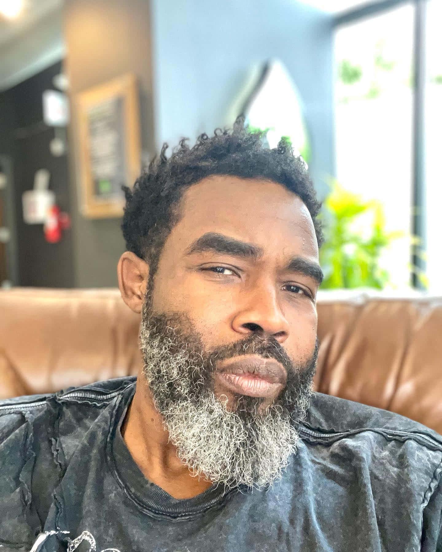  壁紙 — 一位留著灰白鬍鬚和捲髮的男人坐在現代室內環境中的棕色皮革沙發上。