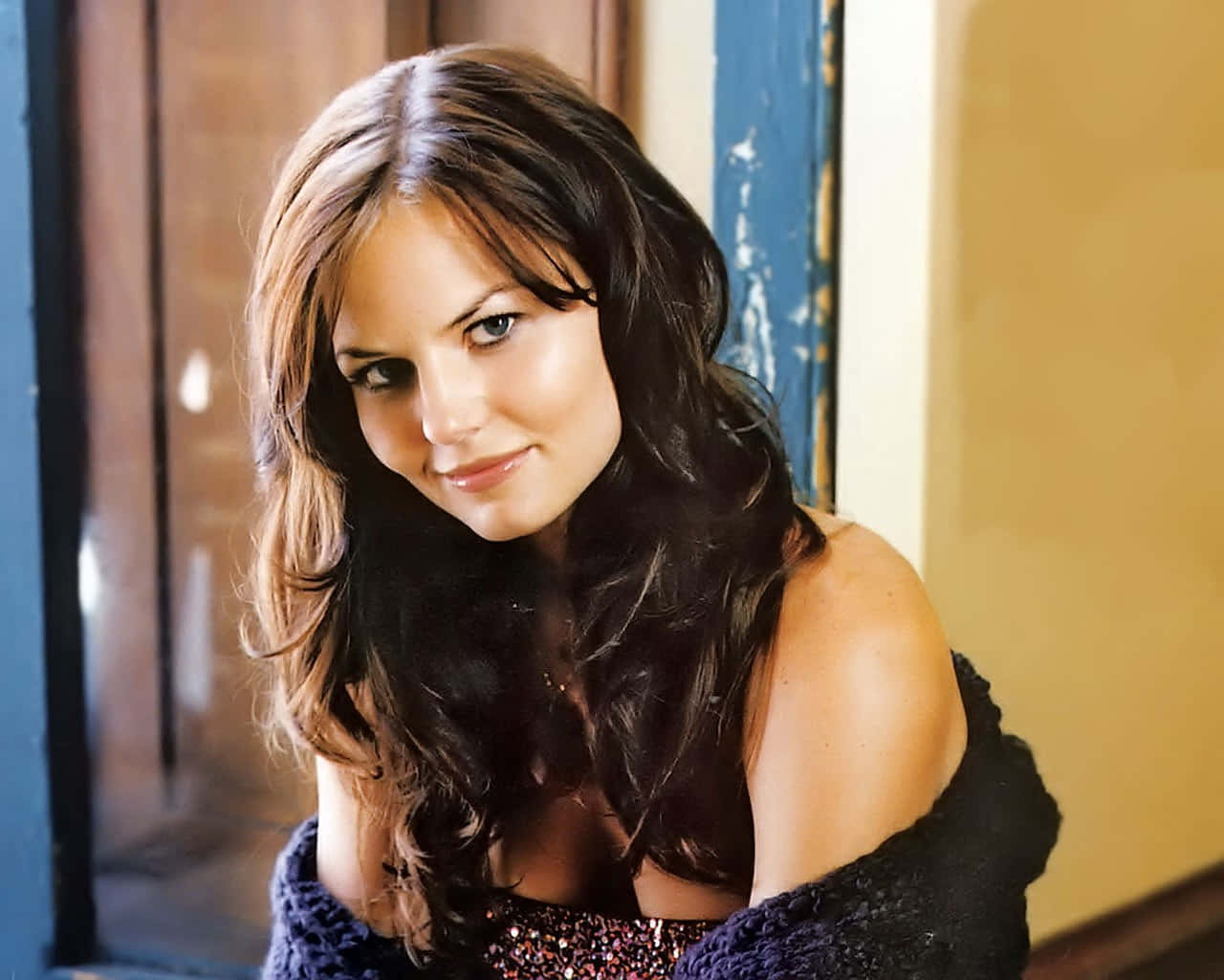 jennifer morrison 桌布 壁紙 — 一位留著深色波浪長髮的女性，輕輕微笑著靠在一個風化的藍色木門框上。