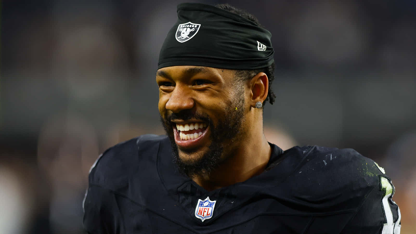  壁紙 — 一位面帶微笑的NFL美式足球員，身穿黑色Raiders球衣和頭帶，在體育場燈光下比賽時開懷大笑。