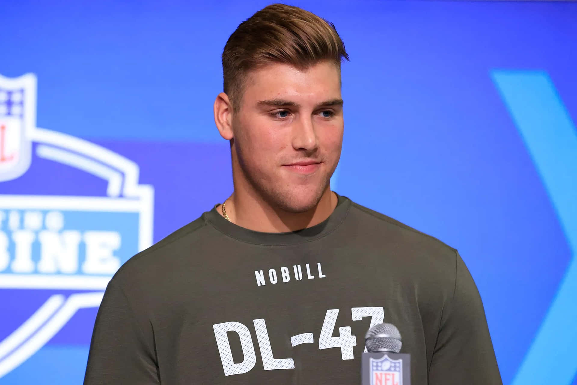  壁紙 — 一位年輕的NFL球員穿著灰色'NOBULL DL-47'襯衫，在藍色品牌背景的新聞發布會上自信地站立。