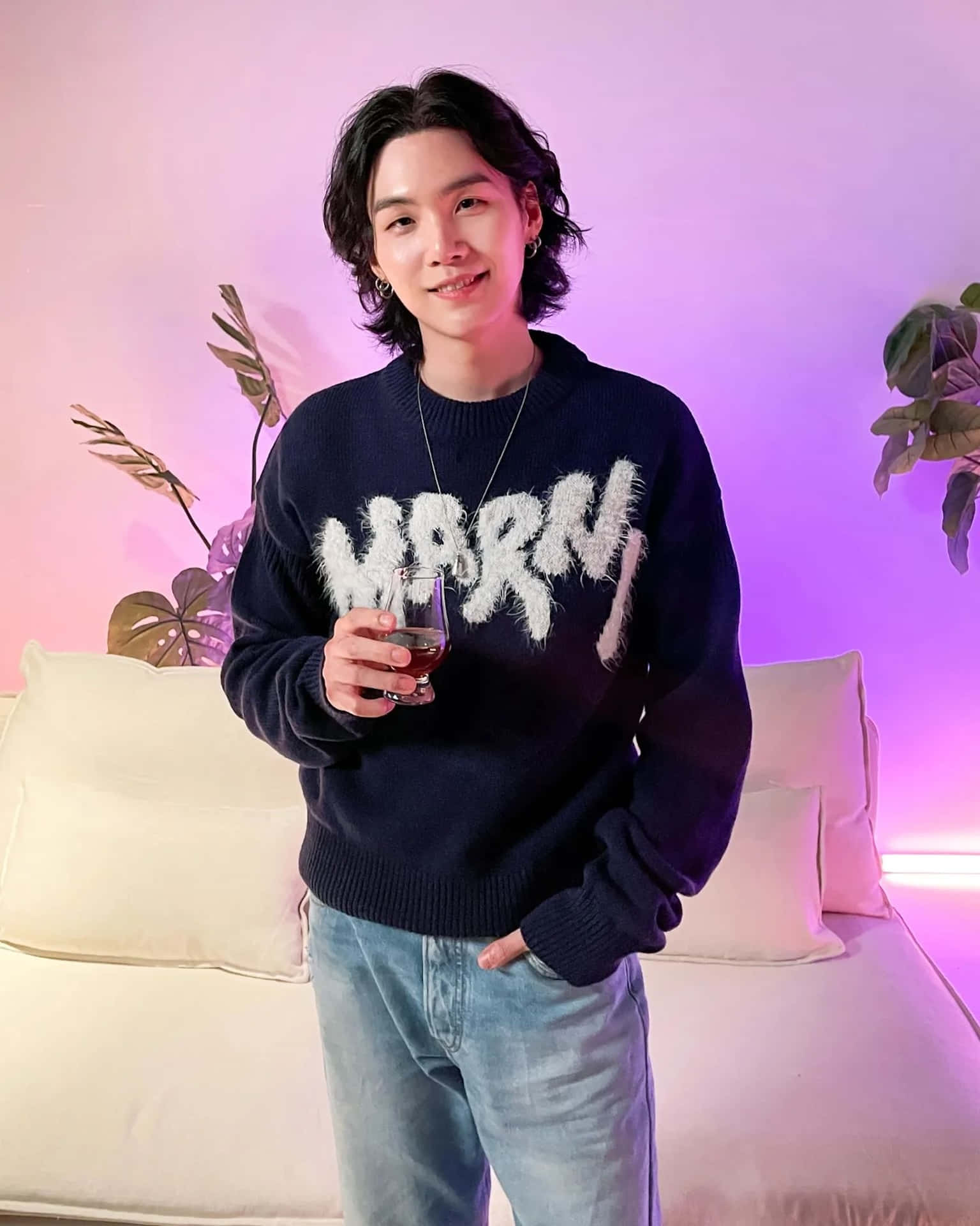  壁紙 — 一位年輕男子留著波浪般的深色頭髮，在一個以粉紅和紫色環境燈光點綴的舒適房間裡，手持一杯深色液體微笑。