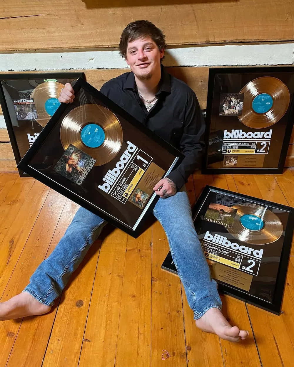  壁紙 — 一位年輕男子微笑著赤腳坐在木地板上，周圍是裱框的Billboard音樂獎項。