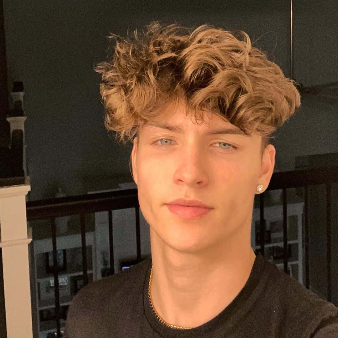  壁紙 — 一位年輕男子，一頭凌亂的金髮和淺色眼睛，在室內柔和燈光下平靜地凝視鏡頭。