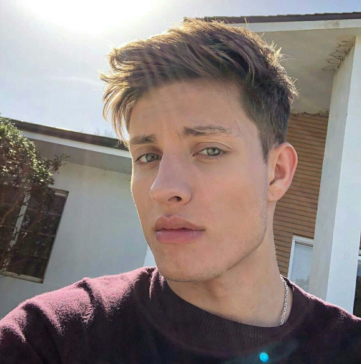 Matt Rife 桌布 壁紙 — 一位年輕男子，棕色頭髮造型整齊，綠色眼睛，在房子附近明亮的陽光下直視鏡頭。