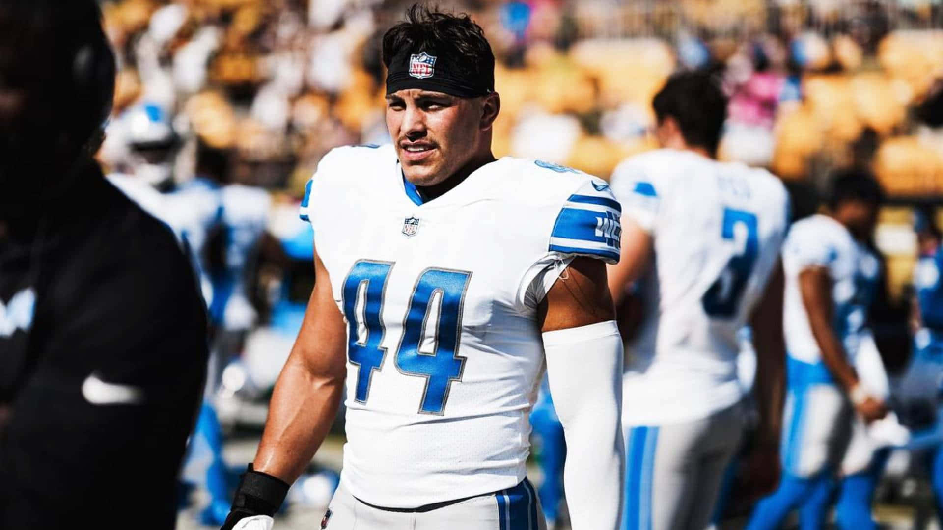 壁紙 — 一位身穿白色和藍色球衣、背號44的專注NFL球員站在陽光明媚的球場上，背景是模糊的隊友和體育場座位。