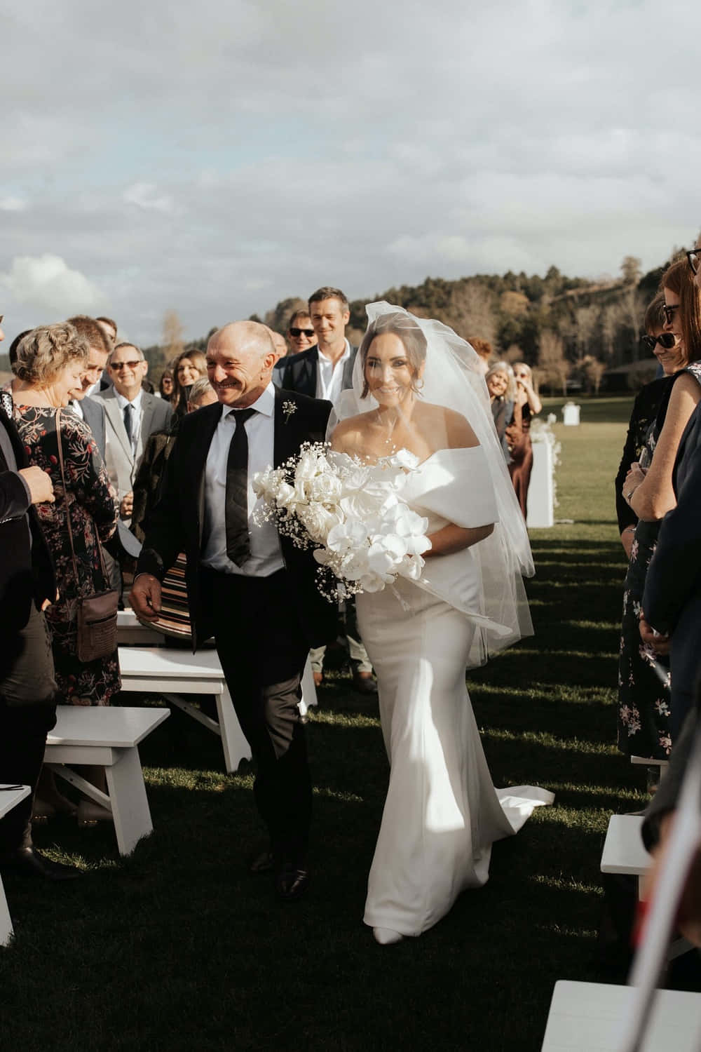 Black And White Wedding 桌布 壁紙 — 一位身穿白色露肩婚紗的新娘，手持白色花束，在草地上走過，父親在她身旁微笑。