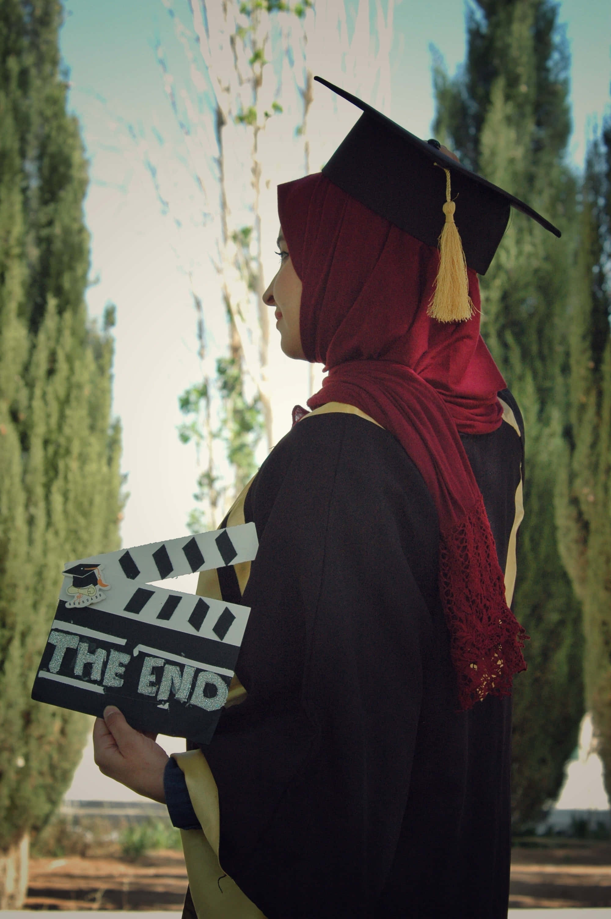 Graduation Ceremony 桌布 壁紙 — 一位身穿黑色畢業袍和學士帽、戴著紅色頭巾的畢業生，在明亮的日光下拿著一塊寫有「THE END」的場記板。